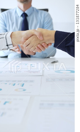 Business people shaking hands in office with financial charts, laptop and reports on table, symbolizing success, teamwork, agreement and corporate growth. Handshake concept 131877284