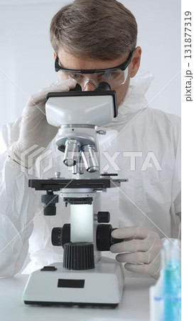A male medical virologist in a medical gown and gloves examines viral samples under a microscope while conducting research in a sterile laboratory setting A male medical virologist in a medical gown and gloves examines viral samples under a microscope while conducting research in a sterile laboratory setting 131877319