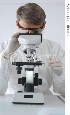 A male medical virologist in a medical gown and gloves examines viral samples under a microscope while conducting research in a sterile laboratory setting A male medical virologist in a medical gown and gloves examines viral samples under a microscope while conducting research in a sterile laboratory setting 131877325