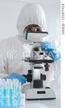 Medical scientist in protective suit using microscope while analyzing samples, conducting research in sterile laboratory environment. Close-up of test tubes with blue liquid Medical scientist in protective suit using microscope while analyzing samples, conducting research in sterile laboratory environment. Close-up of test tubes with blue liquid 131877418