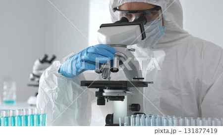 Medical virologist in protective hazmat gear examining viral samples under a microscope while conducting research in a sterile laboratory environment 131877419