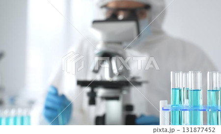 Male medical scientist in protective suit using microscope while analyzing samples, conducting research in laboratory. Close-up of test tubes with blue liquid 131877452