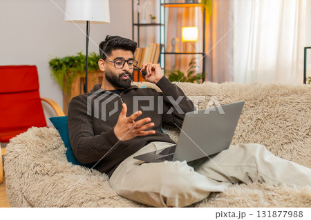 Indian man using wireless headset speaks on laptop video call with client while working at home Indian man using wireless headset speaks on laptop video call with client while working at home 131877988
