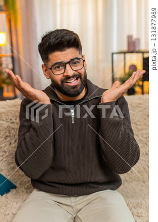 Indian man at sofa spreads hands in confusion and surprise at home, asking what with puzzled face Indian man at sofa spreads hands in confusion and surprise at home, asking what with puzzled face 131877989
