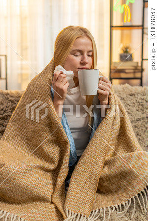 Young woman battling cold at home, wrapped in blanket, drinking hot tea, cozy sofa rest, sniffles Young woman battling cold at home, wrapped in blanket, drinking hot tea, cozy sofa rest, sniffles 131878037