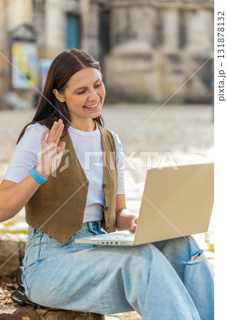 Happy young woman using laptop having video call chat conversation while sitting in city street 131878132