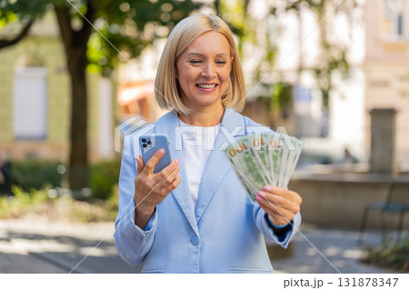 Caucasian mature businesswoman on city street showing earnings cash money with smartphone in hands Caucasian mature businesswoman on city street showing earnings cash money with smartphone in hands 131878347