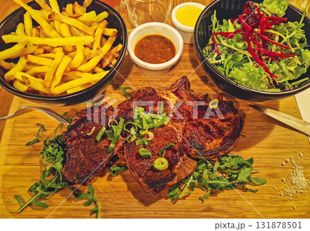 Closeup hearty meal plate with steak chop on a wooden board over arugula, topped with chives and flanked by bowls of golden fries and mixed salad. Small bowls of sauce and mustard complete the 131878501