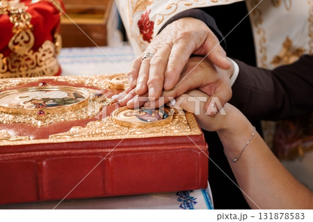 Orthodox Wedding Ceremony: Hands Joined Over Holy Book with Red Cover, Priest's Vestments, Gold Details, Religious Ritual, Faith, Tradition. Orthodox Wedding Ceremony: Hands Joined Over Holy Book with Red Cover, Priest's Vestments, Gold Details, Religious Ritual, Faith, Tradition. 131878583