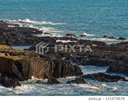 Waves crash against the jagged, dark rocks along the coast at Sheep Cove in Ireland. The sun shines on the water and the irregular stone formations. Waves crash against the jagged, dark rocks along the coast at Sheep Cove in Ireland. The sun shines on the water and the irregular stone formations. 131878676