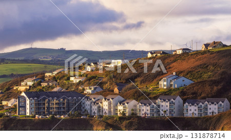 Several houses and buildings are seen on a hillside during the day. Green hills and cloudy skies are in the distance. It looks like a peaceful place to live. 131878714