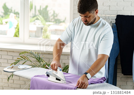 Man ironing a purple shirt in a bright room with plants and clothes in the background Man ironing a purple shirt in a bright room with plants and clothes in the background 131881484