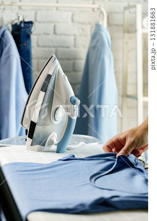 Ironing a blue shirt in a well-lit laundry room filled with neatly organized clothes 131881663