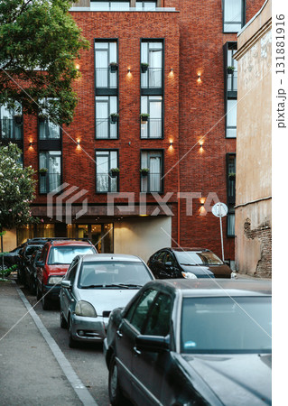 Urban street lined with parked cars near modern brick building in the evening light with greenery Urban street lined with parked cars near modern brick building in the evening light with greenery 131881916