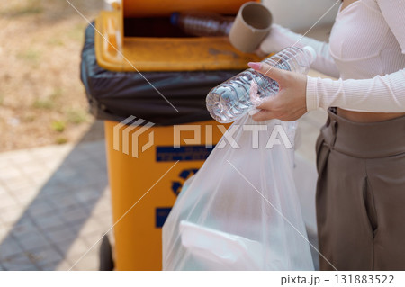 Promoting recycling habits. A woman holds a plastic bottle and a bag while preparing to recycle in an outdoor setting. 131883522