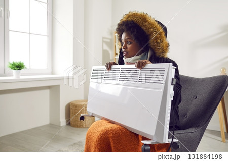 Young pretty african-american woman is sitting in armchair in warm jacket and scarf with heater in her hands in living room, freezing from cold, side view. Young pretty african-american woman is sitting in armchair in warm jacket and scarf with heater in her hands in living room, freezing from cold, side view. 131884198