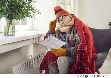Senior man sitting at home near electric heater and looking at the utility bill. 131885747
