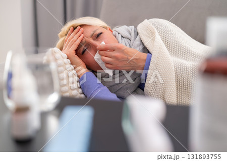 Middle aged blonde woman lying on a couch, covered with a blanket, suffering from a cold and runny nose. Medicines and thermometer on table in foreground. Concept of illness, flu, rest, and home Middle aged blonde woman lying on a couch, covered with a blanket, suffering from a cold and runny nose. Medicines and thermometer on table in foreground. Concept of illness, flu, rest, and home 131893755