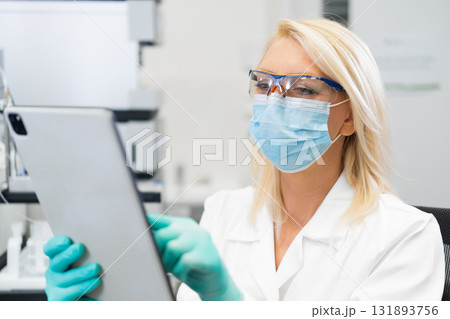 Middle aged female scientist wearing mask and lab coat using tablet in laboratory for research and analysis  131893756