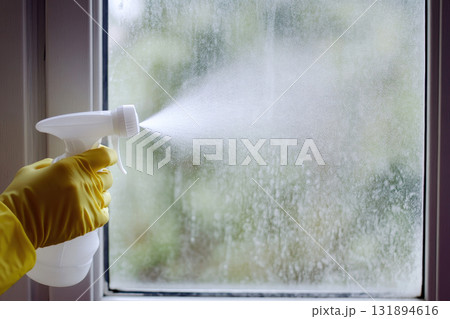 hand in yellow rubber glove holding plastic spray bottle, cleaning detergent on a windows background. 131894616
