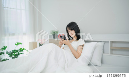 Young woman sitting in cozy bed, using smartphone and enjoying serene morning light in a minimalist bedroom atmosphere with plants and soft bedding 131899676