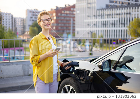 A smiling woman holds her phone while charging her electric vehicle at a charging station in an urban setting. A smiling woman holds her phone while charging her electric vehicle at a charging station in an urban setting. 131902062