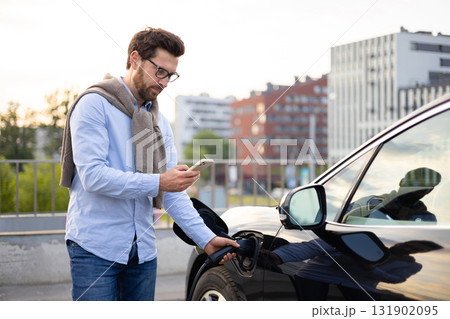 A man in glasses uses his phone while charging his electric car at a charging station in an urban setting. A man in glasses uses his phone while charging his electric car at a charging station in an urban setting. 131902095