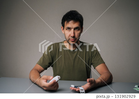 Studio portrait of young man disinfecting smart watch with antibacterial spray cleaner, sitting at table on gray isolated background, taking care of personal hygiene. Concept of healthy lifestyle. Studio portrait of young man disinfecting smart watch with antibacterial spray cleaner, sitting at table on gray isolated background, taking care of personal hygiene. Concept of healthy lifestyle. 131902929