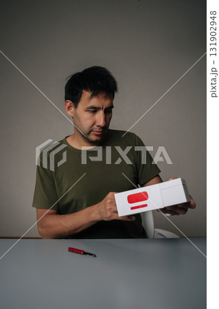 Vertical portrait of skilled blogger male unpacking new electronic cigarette using small knife sitting at desk on isolated gray background. Concept of modern alternative to traditional smoking. 131902948