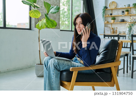 Positive young woman working at home with laptop talking on phone Positive young woman working at home with laptop talking on phone 131904750
