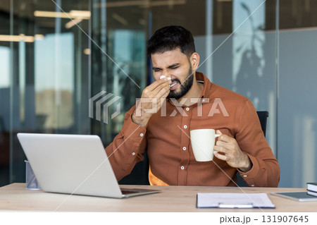 Young professional at office desk battling cold or allergies, holding warm cup and tissue while working on his computer, showing fatigue and discomfort during workday 131907645