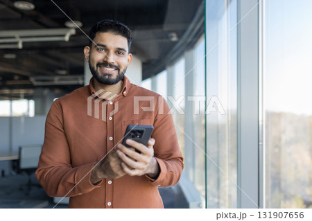 Indian businessman smiling at camera, holding a smartphone near a large window in a contemporary high-rise office, representing communication, technology, and success Indian businessman smiling at camera, holding a smartphone near a large window in a contemporary high-rise office, representing communication, technology, and success 131907656