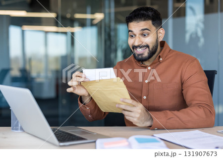 Excited young professional man sitting at his office desk opening a brown envelope with a surprised happy expression, learning about a promotion or financial success 131907673