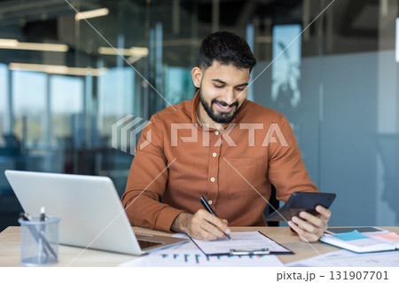 Indian businessman smiling at desk while reviewing paperwork with laptop and calculator, managing budgeting, taxes and finances in a modern office environment Indian businessman smiling at desk while reviewing paperwork with laptop and calculator, managing budgeting, taxes and finances in a modern office environment 131907701
