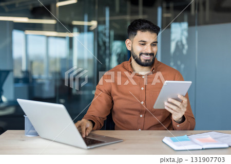 Young businessman happily working at office desk, holding a digital tablet while typing on a laptop, demonstrating efficiency, connectivity, and modern technology integration in daily tasks Young businessman happily working at office desk, holding a digital tablet while typing on a laptop, demonstrating efficiency, connectivity, and modern technology integration in daily tasks 131907703