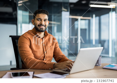 Indian businessman sitting at an office desk, smiling while working on a laptop, symbolizing professionalism, success, and a positive corporate environment in a modern workspace 131907741
