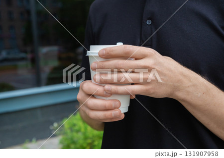 man holds disposable coffee cup with both hands and warms his hands on hot mug. coffee break. White paper kraft disposable cup for coffee with white plastic lid. Take-away cup. man holds disposable coffee cup with both hands and warms his hands on hot mug. coffee break. White paper kraft disposable cup for coffee with white plastic lid. Take-away cup. 131907958