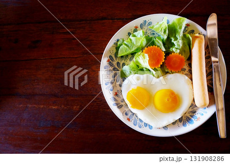 Fresh heart shaped fried eggs with organic vegetable salad and hot dog in white dish breakfast on wooden table Fresh heart shaped fried eggs with organic vegetable salad and hot dog in white dish breakfast on wooden table 131908286