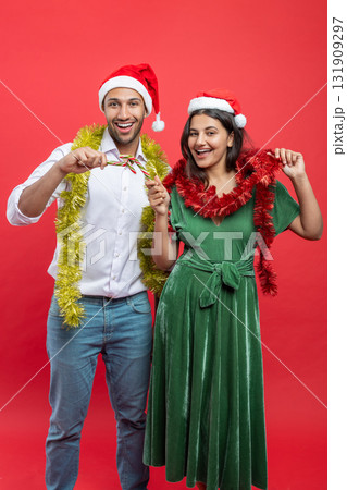 Happy couple dressed in Santa hats and tinsel celebrating new year together holding candy canes Happy couple dressed in Santa hats and tinsel celebrating new year together holding candy canes 131909297
