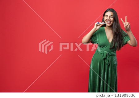 Playful woman enjoying her Christmas celebration indoors blowing noisemaker showing v-sign 131909336