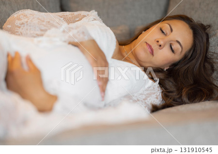 Pregnant woman resting serenely on couch wearing elegant white lace attire Pregnant woman resting serenely on couch wearing elegant white lace attire 131910545