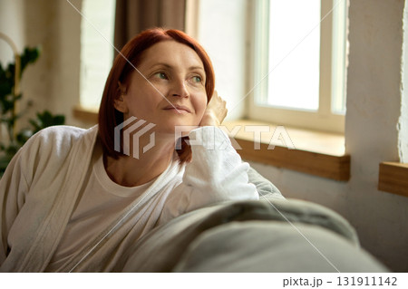 Adult woman resting on sofa near window with gentle daylight and peaceful thoughtful expression 131911142