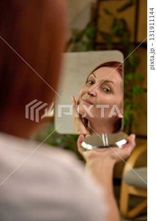 Mature woman gently touching face while admiring reflection in mirror with soft smile 131911144