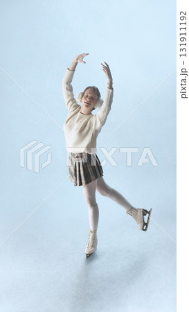 Smiling female skater performing joyful pose on ice in cozy winter outfit under soft light 131911192
