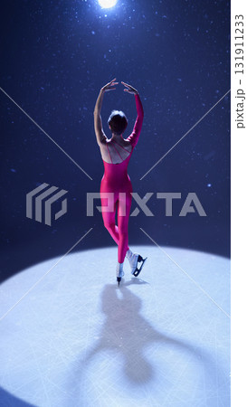 Woman figure skater in pink costume raising arms to light in elegant ballet-inspired position Woman figure skater in pink costume raising arms to light in elegant ballet-inspired position 131911233
