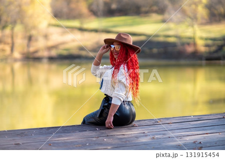 Autumn lake woman. In autumn, she sits by the pond on a wooden pier and admires nature with red hair and a hat. Tourism concept, weekend outside the city. Autumn lake woman. In autumn, she sits by the pond on a wooden pier and admires nature with red hair and a hat. Tourism concept, weekend outside the city. 131915454