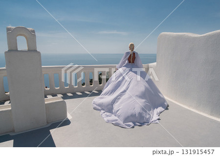 Santorini Bride Dress: Woman in white dress observes Aegean Sea from the whitewashed balcony under daylight. Santorini Bride Dress: Woman in white dress observes Aegean Sea from the whitewashed balcony under daylight. 131915457