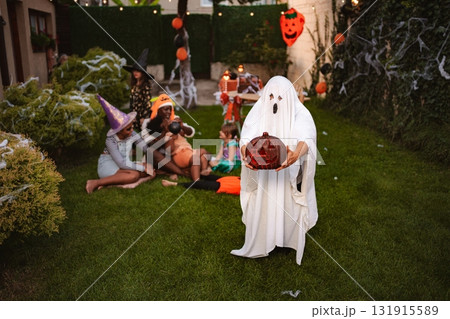 Child wearing ghost costume holding jack-o'-lantern during Halloween party in the garden Child wearing ghost costume holding jack-o'-lantern during Halloween party in the garden 131915589