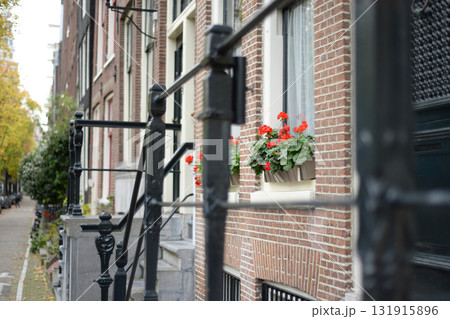 A close-up view of a brick building in Amsterdam. The facade features a black wrought iron railing and flower boxes with red geraniums. Trees line the street A close-up view of a brick building in Amsterdam. The facade features a black wrought iron railing and flower boxes with red geraniums. Trees line the street 131915896