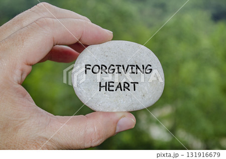Hand holding a white stone with FORGIVING HEART text engraved on it, symbolizing life purpose of forgiveness and emotional peace Hand holding a white stone with FORGIVING HEART text engraved on it, symbolizing life purpose of forgiveness and emotional peace 131916679